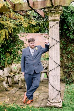 A man in a suit leans against a vine-wrapped column in a garden setting, reminiscent of an Elmhay Park Wedding. Stone benches and lush greenery provide the perfect picturesque backdrop.