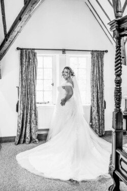 The bride, captured by an Elmhay Park wedding photographer, stands smiling by a window in a room with exposed wooden beams and curtained windows, her long, flowing wedding dress gracefully cascading around her.