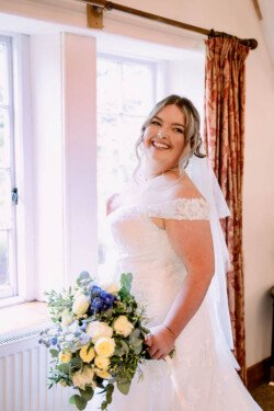 Evoto Bride smiling near a window at Elmhay Park, holding a bouquet with white and blue flowers, in a white wedding dress with lace details.