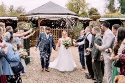 Bride and groom walk down the aisle beneath the sky as guests shower them with confetti, all set against the enchanting backdrop captured by Elmhay Park Wedding Photographer. A picturesque canopy structure adds to the perfect outdoor setting.