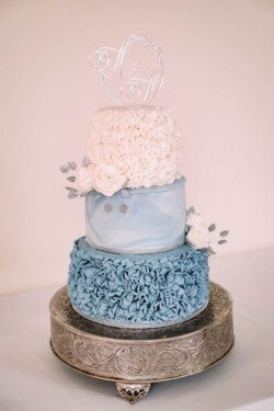 A stunning three-tiered cake with blue and white icing, floral decorations, and a monogram topper is beautifully showcased on an ornate metal stand—perfect for capturing memorable moments as an Elmhay Park wedding photographer.