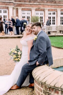 Bride and groom sit together on a brick structure outdoors, with the bride holding a bouquet and smiling. Guests mingle in the background as an Elmhay Park Wedding Photographer captures the joyful scene.