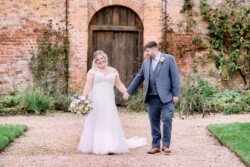 Bride and groom holding hands and smiling in front of a rustic brick wall, with the bride holding a bouquet, beautifully captured by an Elmhay Park wedding photographer.