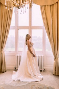 Evoto The bride, in a white gown and veil, stands gracefully by tall windows with beige curtains at Orchardleigh House Weddings, under a dazzling chandelier.
