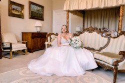 Evoto A bride in a white gown sits on an ornate sofa, holding a bouquet, in a well-decorated room at Orchardleigh House Weddings.