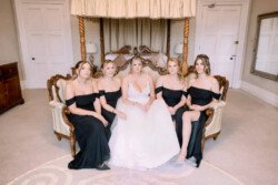Evoto In the elegant setting of Orchardleigh House Weddings, a bride in a white gown sits gracefully on a vintage sofa alongside four bridesmaids in black dresses, all surrounded by the charm of an exquisite room featuring a lovely canopy bed.
