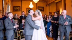 Amidst the elegance of Orchardleigh House Weddings, a bride and groom share a kiss at their ceremony, surrounded by guests who are clapping and smiling.