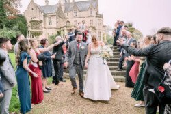 Evoto A newlywed couple walks down steps outside the historic Orchardleigh House, greeted by cheering guests.