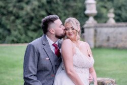 Evoto A man in a suit kisses a smiling woman in a white dress. They stand outdoors near a stone wall, surrounded by the lush greenery typical of Orchardleigh House Weddings.