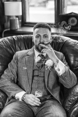 A man in a suit sits in a leather chair, exuding an air of refined elegance as he holds a cigar and a glass with a drink, reminiscent of the sophisticated ambiance at Orchardleigh House weddings.