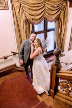 Evoto At Orchardleigh House Weddings, a couple in formal attire poses gracefully on a wooden staircase adorned with ornamental drapery and overlooking a large window. The woman is elegantly dressed in white, while the man complements her charm in a sleek suit.