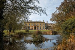 A large historical manor house, often sought by couples for its enchanting backdrop, features a pond in the foreground and is surrounded by lush trees and greenery. Ideal for capturing timeless moments, this serene setting is a dream for any Hartham Park wedding photographer.