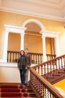 Two people stand on a grand staircase with ornate railings, captured perfectly by a Hartham Park wedding photographer, in a building with yellow walls and archways.
