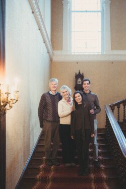 Four people stand on a staircase in a well-lit room with a large window, captured by our Hartham Park wedding photographer. Two are older adults, and two are younger adults. They are smiling and casually dressed, enjoying the perfect blend of light and moment.