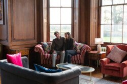 A couple sits on a striped couch in a wood-paneled room with large windows and colorful cushions, their expressions captured beautifully by a Hartham Park wedding photographer.