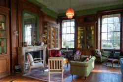 A cozy library room with large windows and wooden shelves filled with books invites you to unwind. The fireplace crackles nearby, while a variety of chairs and sofas offer comfort as you enjoy the landscape view—the perfect backdrop for a Hartham Park Wedding Photographer.