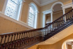 In the historic setting of Hartham Park, a grand wooden staircase with ornate balusters ascends gracefully. Stained glass windows illuminate the space, enhanced by classical architectural details and cream-colored walls, providing a stunning backdrop ideal for any wedding photographer.