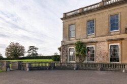 A large stone building with tall windows and ivy, perfect for a Hartham Park wedding photographer, stands adjacent to a paved area and a grassy field, framed by trees under a cloudy sky.