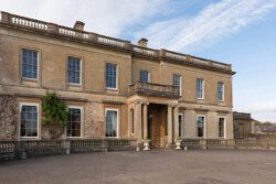 A large, historic stone mansion with ivy-covered walls and a reputation among Hartham Park wedding photographers, it showcases two stories, multiple windows, and a central entrance framed by columns under a clear blue sky.