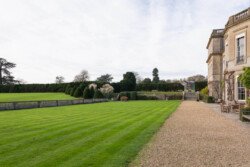 A manicured lawn with a gravel path leads to an elegant, large stone building, perfect for capturing unforgettable moments. Shrubs line the horizon under a partly cloudy sky, making it an ideal setting for a Hartham Park wedding photographer.