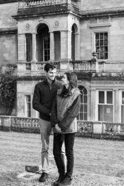A couple stands in front of a historic building, smiling at each other as if captured by a Hartham Park Wedding Photographer. The structure boasts large windows and ornate stonework, providing the perfect backdrop for their special moment.
