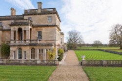 A grand stone mansion, ideal for a Hartham Park wedding photographer, boasts multiple levels and a columned entrance. It's surrounded by a well-maintained lawn and gravel path, set against a backdrop of trees and a partly cloudy sky.