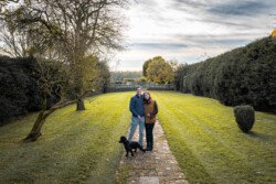 A couple stands on a stone path in a garden with their black dog, perfectly captured by a Hartham Park Wedding Photographer. The sky is partly cloudy, and trees and hedges line the lush grass.