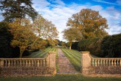 A stone balustrade frames a pathway lined with trees, leading towards a distant garden under the blue sky with white clouds. The trees display autumn foliage—a picturesque scene perfect for capturing by a Hartham Park Wedding Photographer.