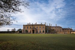 The large historic mansion, perfect for a Hartham Park wedding photographer, boasts multiple chimneys and is surrounded by a grassy lawn and trees under a partly cloudy sky.
