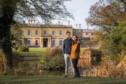 Two people stand in front of a large historic building at Hartham Park, surrounded by trees and a small pond, capturing the essence of romance in this enchanting setting, perfect for any wedding photographer.
