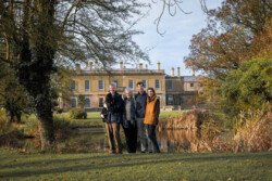 Four people stand together on a grassy area in front of a large, yellow mansion surrounded by trees and a pond, the scene beautifully captured by a Hartham Park wedding photographer.
