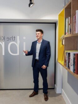 OLYMPUS DIGITAL CAMERA A man in a blue suit stands in a modern office, gesturing with his hand. Shelves with books are visible on the right, and a frosted glass door with partial text is behind him. The scene captures the essence of professional Corporate Photography & Video Services.