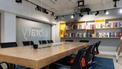 OLYMPUS DIGITAL CAMERA A modern conference room with a large wooden table, black chairs, and bookshelves filled with books on the back wall. The word "Library" is visible on a glass panel, perfectly staged for corporate photography & video services.