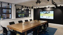 OLYMPUS DIGITAL CAMERA A modern conference room with a large wooden table, black chairs, a wall-mounted screen displaying a presentation, and multiple windows—perfectly capturing the essence of Corporate Photography & Video Services.