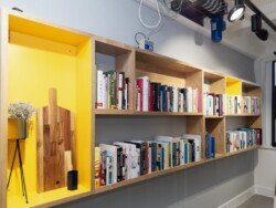 OLYMPUS DIGITAL CAMERA A wooden bookshelf with multiple compartments filled with various books. The leftmost section contains a decorative plant, a cutting board, and a small blue container—ideal for a touch of personality in corporate photography & video services setups.