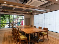 OLYMPUS DIGITAL CAMERA A modern meeting room with a large wooden table surrounded by chairs. One wall features a large window with a view of an outdoor seating area and greenery, perfect for corporate photography & video services.