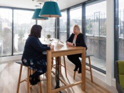 OLYMPUS DIGITAL CAMERA Two women sit at a high wooden table in a bright room with large windows and teal hanging lamps, engaged in conversation—a perfect scene for corporate photography & video services.