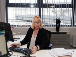 OLYMPUS DIGITAL CAMERA A woman sits at an office desk working on a computer, surrounded by paperwork and office equipment, with a window and flower pot in the background, perfectly capturing the essence of corporate photography.