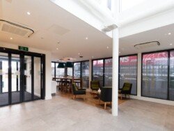 OLYMPUS DIGITAL CAMERA A modern office lobby with glass doors, seating area with armchairs and a wooden table, and windows with black blinds. The floor is a mix of tile and wooden panels, perfect for showcasing your space through Corporate Photography & Video Services.