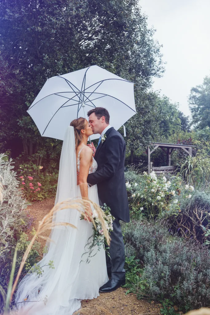 A bride and groom kiss under an umbrella in a garden. Weddings in bath