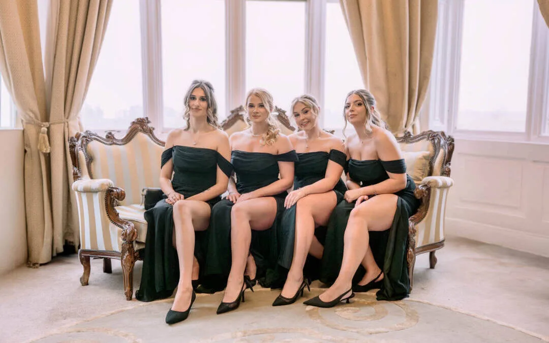 Four women in black dresses sit on a vintage sofa in a well-lit room at Orchardleigh House Weddings, with large windows and elegant curtains.