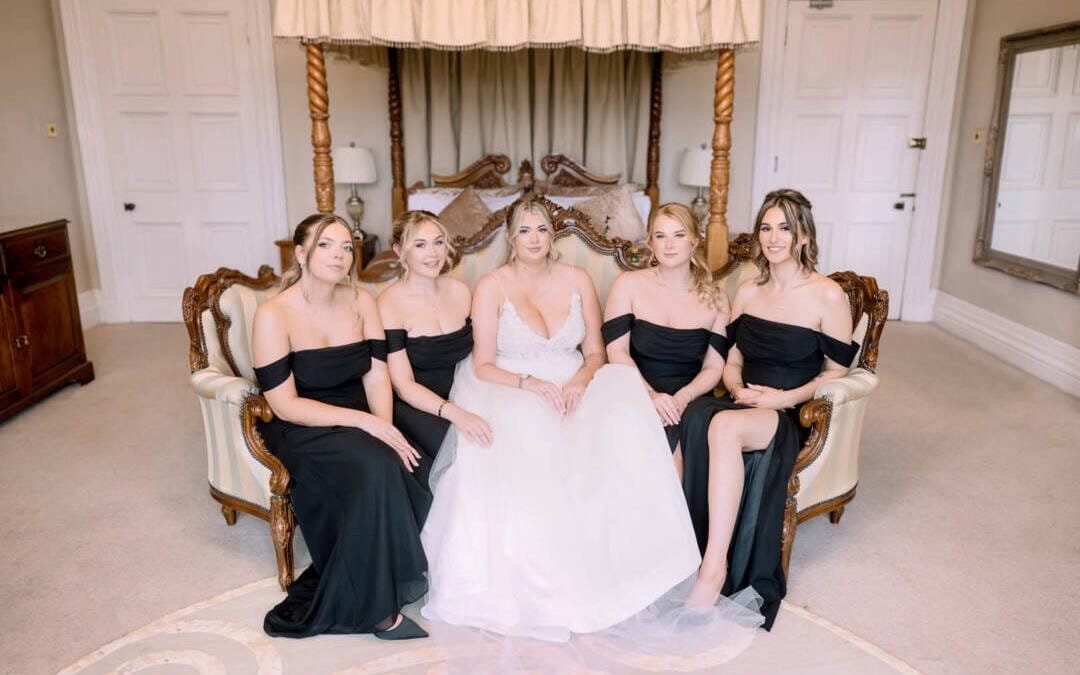 In the elegant setting of Orchardleigh House Weddings, a bride in a white gown sits gracefully on a vintage sofa alongside four bridesmaids in black dresses, all surrounded by the charm of an exquisite room featuring a lovely canopy bed.