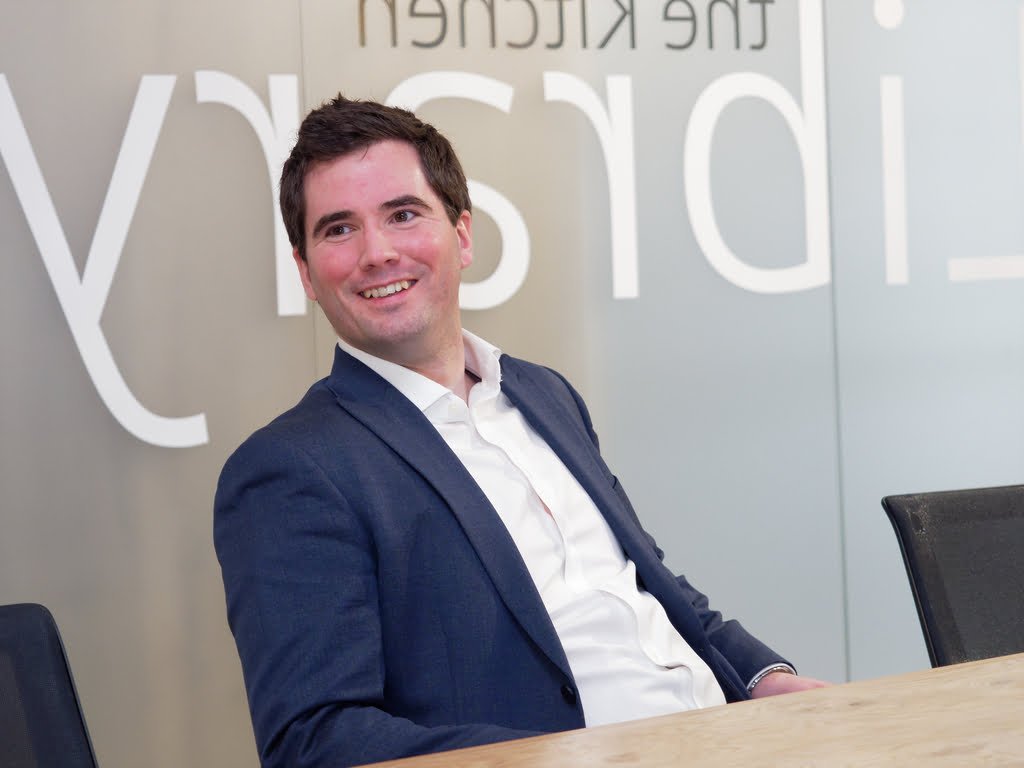 A man wearing a suit and white shirt is sitting at a table, smiling. Partial text from a wall sign in the background subtly hints at Corporate Photography & Video Services.