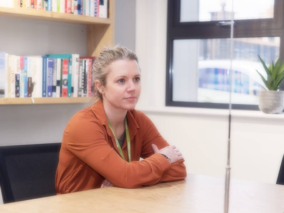 A woman with blonde hair and an orange shirt sits at a wooden table with her arms crossed, in an office setting featuring a bookshelf and a window. This image showcases our Corporate Photography & Video Services, capturing professional environments with precision.