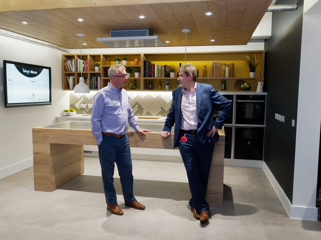 Two men converse in a modern kitchen setting. One leans against the counter, while the other stands with hands in pockets. The kitchen features a wooden ceiling, sleek shelves with books, and a large monitor—an ideal backdrop for corporate photography & video services.