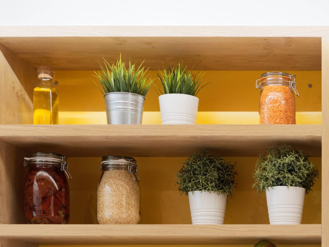 A wooden shelving unit displays potted plants, a bottle of oil, red lentils, dried tomatoes, and grains in glass jars against a yellow backdrop, creating an inviting scene perfect for showcasing your corporate photography & video services.