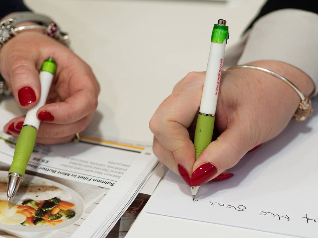 Two hands with red nail polish holding green and white pens; one hand is writing on a sheet of paper while the other holds another pen above a stack of pamphlets or magazines, perfectly capturing a scene ideal for showcasing Corporate Photography & Video Services.