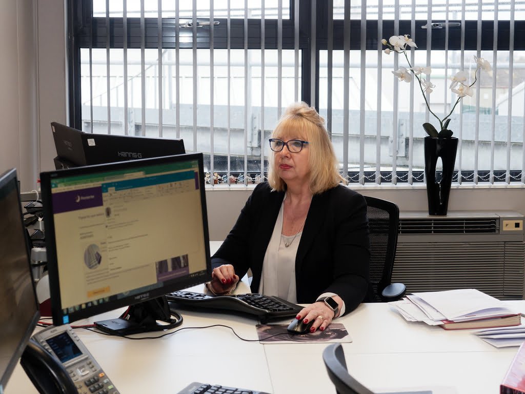 A woman with blonde hair wearing glasses and a dark blazer is seated at a desk with multiple computer monitors in a well-lit office, showcasing the professional environment perfect for Corporate Photography & Video Services. There is a potted orchid and various papers on her desk.