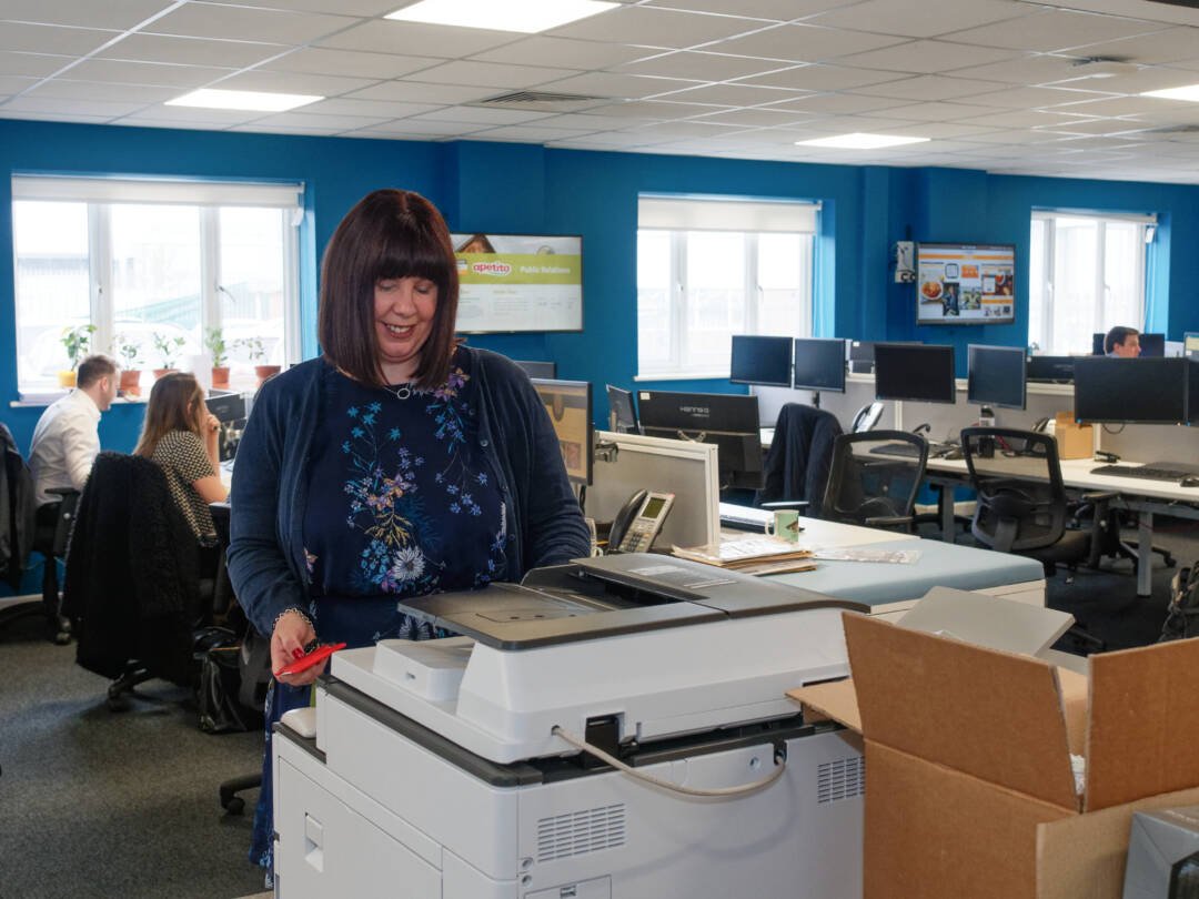 A woman uses a photocopier in an office with several desks and computers, perfectly captured through our Corporate Photography & Video Services. Other employees work at their desks in the background, showcasing a dynamic and productive workplace.